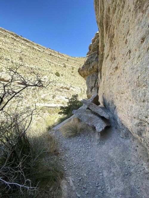 A narrow trail winds between rocky cliffs in a desert landscape under a clear blue sky.
