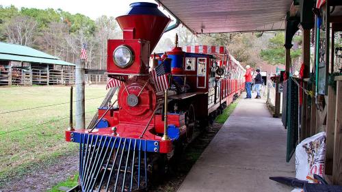 A colorful vintage train with flags parked beside a building, surrounded by greenery and people in the background.