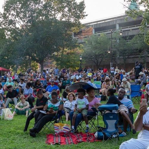 A large crowd sits on lawn chairs in a park, enjoying an outdoor event on a sunny day. Trees and buildings in the background.