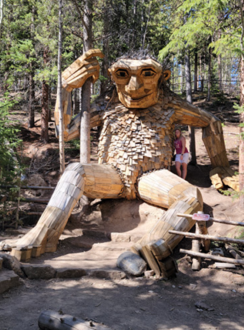 A large wooden troll sculpture sits among trees, with a person standing beside it for scale.