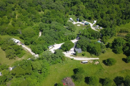 Aerial view of a lush green landscape with winding paths, buildings, and camping areas surrounded by trees.