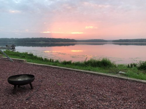 A tranquil lake at sunrise, reflecting soft pink and orange hues, with a fire pit in the foreground and lush greenery.