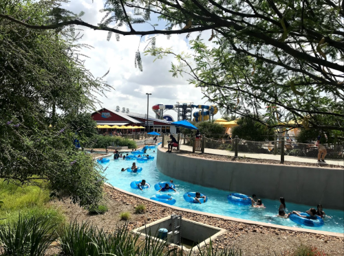 A lazy river at a water park with people floating on inner tubes, surrounded by greenery and water slides in the background.