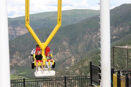 Two children sit in a yellow amusement park ride, suspended high above a green mountainous landscape.
