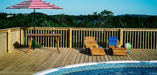 A wooden deck with two lounge chairs, an umbrella, and a pool, surrounded by greenery and a clear blue sky.