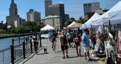 A bustling waterfront market with people walking, tents set up, and city buildings in the background on a sunny day.