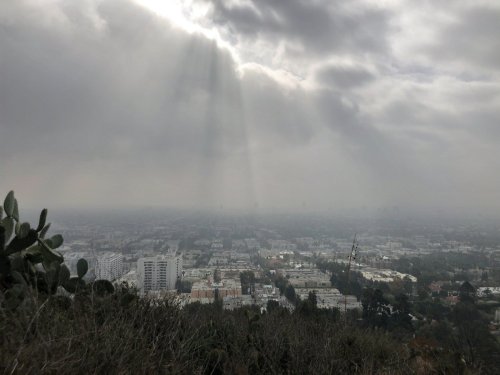 A hazy cityscape under cloudy skies, with sunlight breaking through the clouds and illuminating parts of the urban landscape.