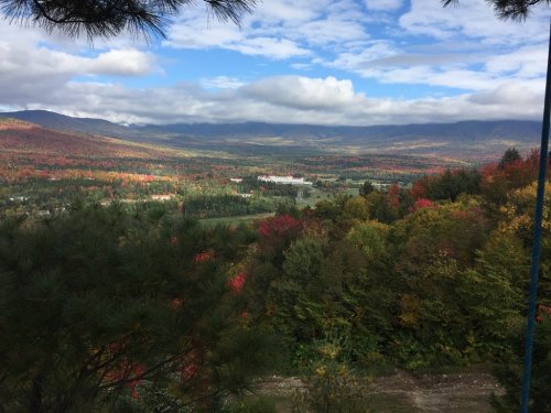 A scenic view of a valley with colorful autumn foliage, mountains in the background, and a partly cloudy sky.