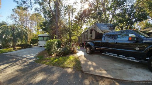 Two RVs parked in a shaded area surrounded by trees and greenery, with a black truck parked beside one of them.