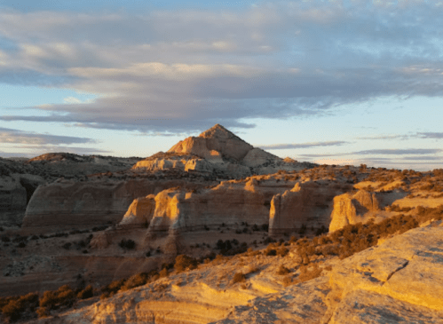 Sunset over colorful rock formations and a mountain peak, with clouds in a blue sky.