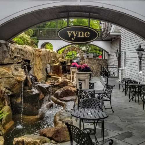 A charming outdoor seating area with a stone waterfall, surrounded by greenery and a sign for "vyne."