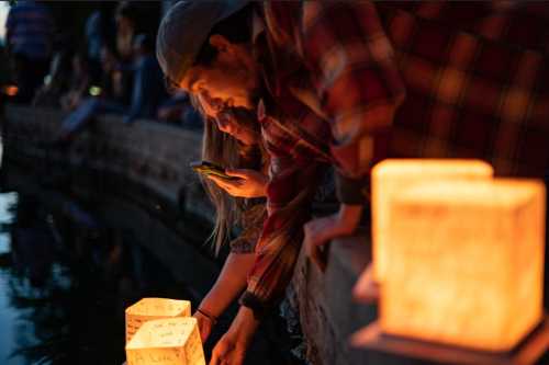 Two people gently placing glowing lanterns into a waterway at dusk, surrounded by a crowd enjoying the moment.