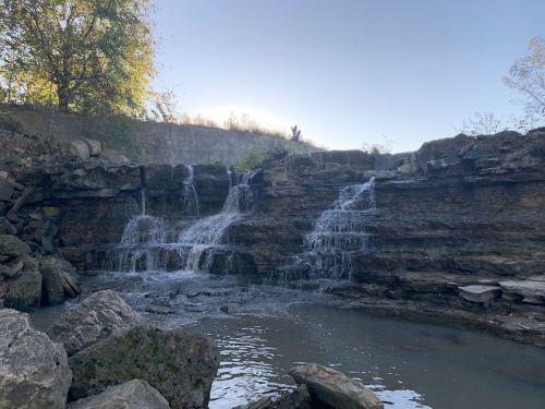 A serene waterfall cascading over rocky ledges into a calm pool, surrounded by trees and a clear sky.