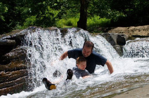 A man and a boy play in a waterfall, splashing water and enjoying a sunny day outdoors.