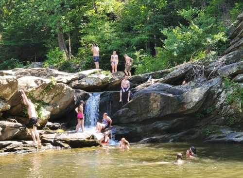 A group of people enjoying a sunny day by a rocky river, swimming and climbing near a small waterfall.