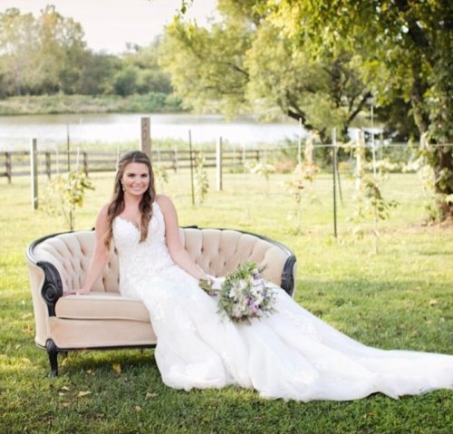 A bride in a white gown sits on a vintage couch in a lush outdoor setting, holding a bouquet of flowers.