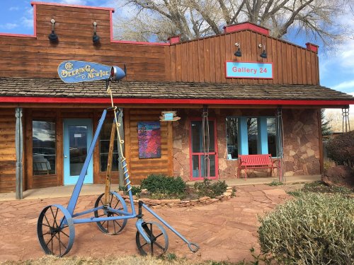 A rustic wooden building with a sign for "Gallery 24" and a blue sculpture in front on a stone pathway.