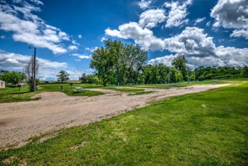 A scenic view of a grassy area with a dirt path, trees, and a bright blue sky filled with fluffy clouds.