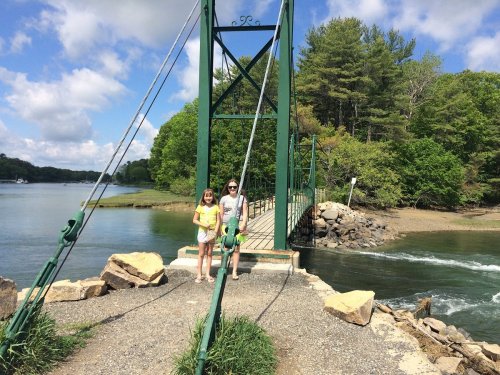 Two people stand on a bridge over a river, surrounded by trees and a partly cloudy sky.