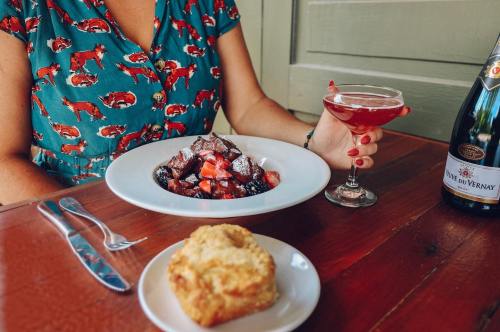A person in a patterned dress holds a cocktail beside a plate of mixed berries and a biscuit on a wooden table.