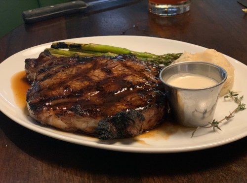A grilled steak on a plate with asparagus, mashed potatoes, and a small cup of sauce.