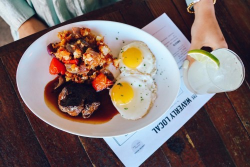 A plate of breakfast featuring two eggs, hash browns, and a piece of meat, alongside a drink with a lime wedge.