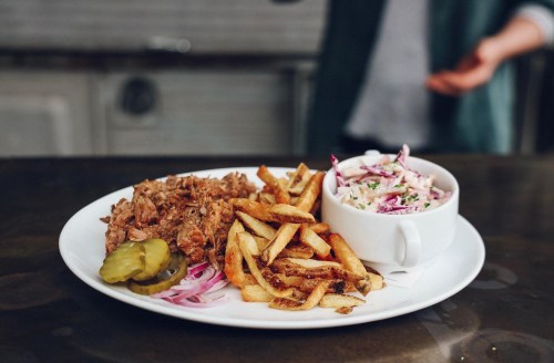 A plate with pulled pork, pickles, fries, and a side of coleslaw on a dark table.