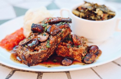 A plate of meatloaf topped with mushrooms, served with mashed potatoes, collard greens, and a side of salsa.