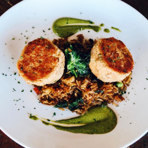 Two golden-brown patties on a bed of rice with greens, drizzled with green sauce, garnished with herbs.