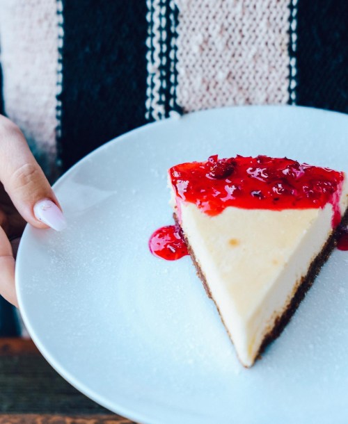 A slice of cheesecake topped with red fruit sauce on a white plate, held by a hand.