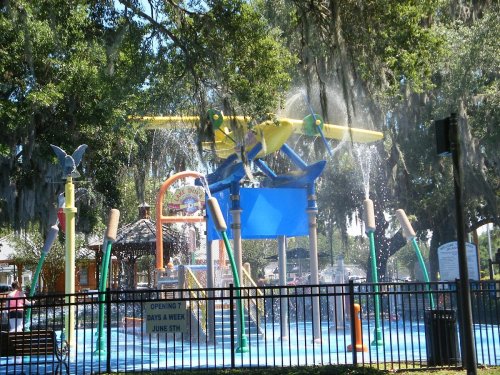 Colorful water play area featuring a yellow and green airplane spraying water, surrounded by trees and a fence.