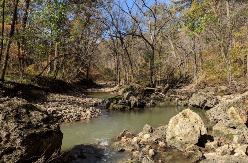 A serene river flows through a rocky landscape, surrounded by trees with autumn foliage under a clear blue sky.