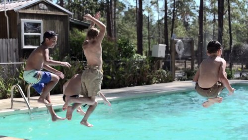 Four children are jumping into a pool on a sunny day, splashing water around in a fun summer scene.