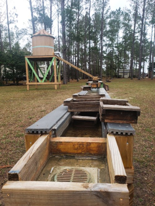 A wooden water trough system with a water tower in the background, surrounded by trees in a grassy area.