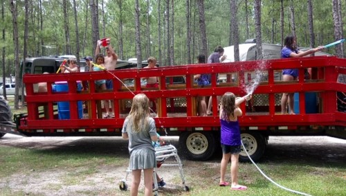 A group of children playfully spraying water from a trailer in a wooded area, enjoying a fun summer day.