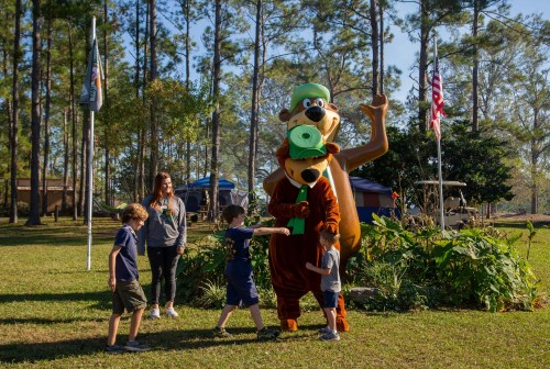 A costumed bear character poses with children in a grassy area surrounded by trees and flags.