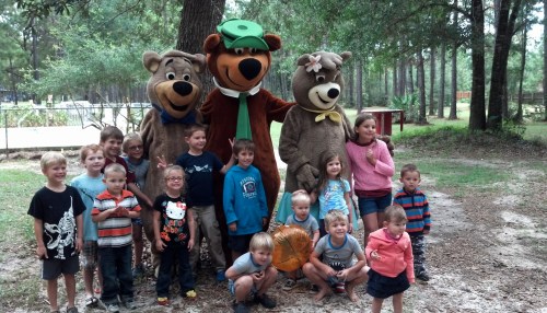 A group of children poses with two bear mascots in a wooded area, smiling and making playful gestures.