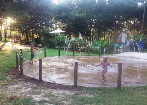 Two children play in a splash pad, enjoying water sprays in a sunny park surrounded by trees.