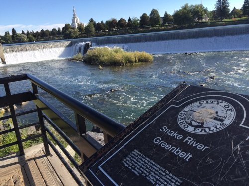 View of the Snake River with a waterfall, a sign about the Greenbelt, and a distant building in Idaho Falls.