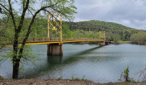 A yellow suspension bridge spans a calm river, surrounded by lush green trees and rolling hills under a cloudy sky.