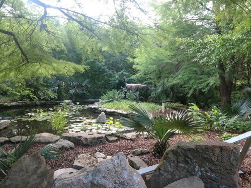 A lush garden with a pond, surrounded by greenery and rocks, featuring a dinosaur statue in the background.