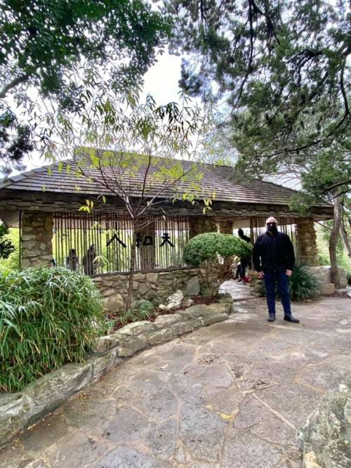 A stone building with a gated entrance surrounded by greenery, with a person standing nearby on a pathway.