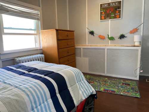 A cozy bedroom with a striped bedspread, wooden dresser, and dinosaur-themed decor on the walls.