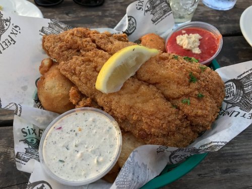 A plate of fried fish fillets with a lemon wedge, served with dipping sauces and hushpuppies on a paper-lined tray.