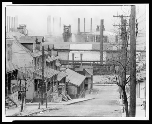 Black and white image of a street lined with houses, with industrial smokestacks and factories in the background.