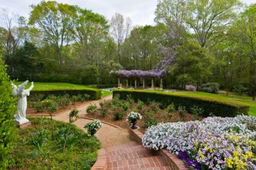 A serene garden path lined with flowers, leading to a pergola adorned with purple blooms and surrounded by lush greenery.