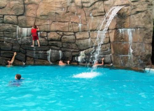 Children playing in a pool with a rock wall and waterfall, one climbing while others swim nearby.