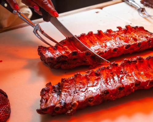 A close-up of a hand using a carving fork and knife to slice into juicy, glazed ribs on a cutting board.
