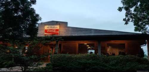 A rustic restaurant with a Coca-Cola sign, surrounded by greenery, under a cloudy sky. Warm lights glow from the windows.