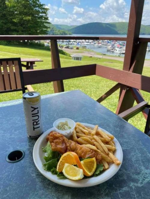 A plate of fried fish, fries, and orange slices, with a can of Truly, set against a scenic lakeside view.